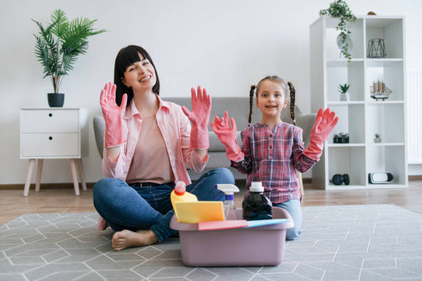 Mother and daughter sit on the floor wearing pink cleaning gloves, ready to clean, with supplies nearby.